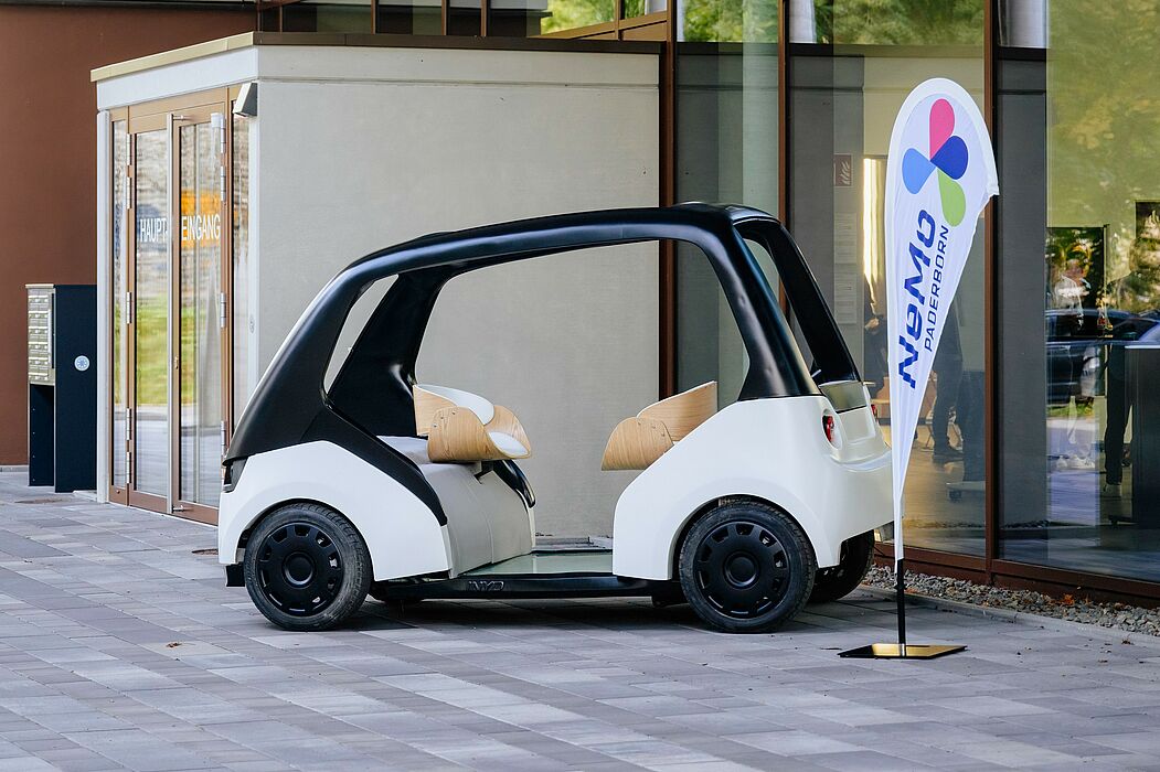 In the foreground, a so-called "Cab", a model vehicle with which people are to be sustainably mobile in the "New Mobility Paderborn" project. Next to it is a flag with the project logo.